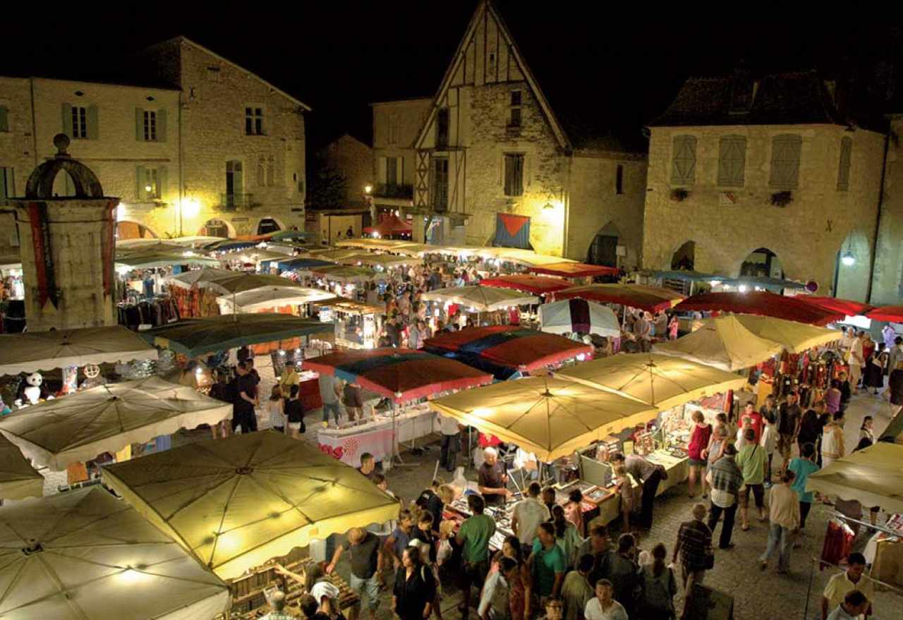 Marché nocturne animé sur une place historique éclairée, avec des stands sous parasols et une foule de visiteurs.